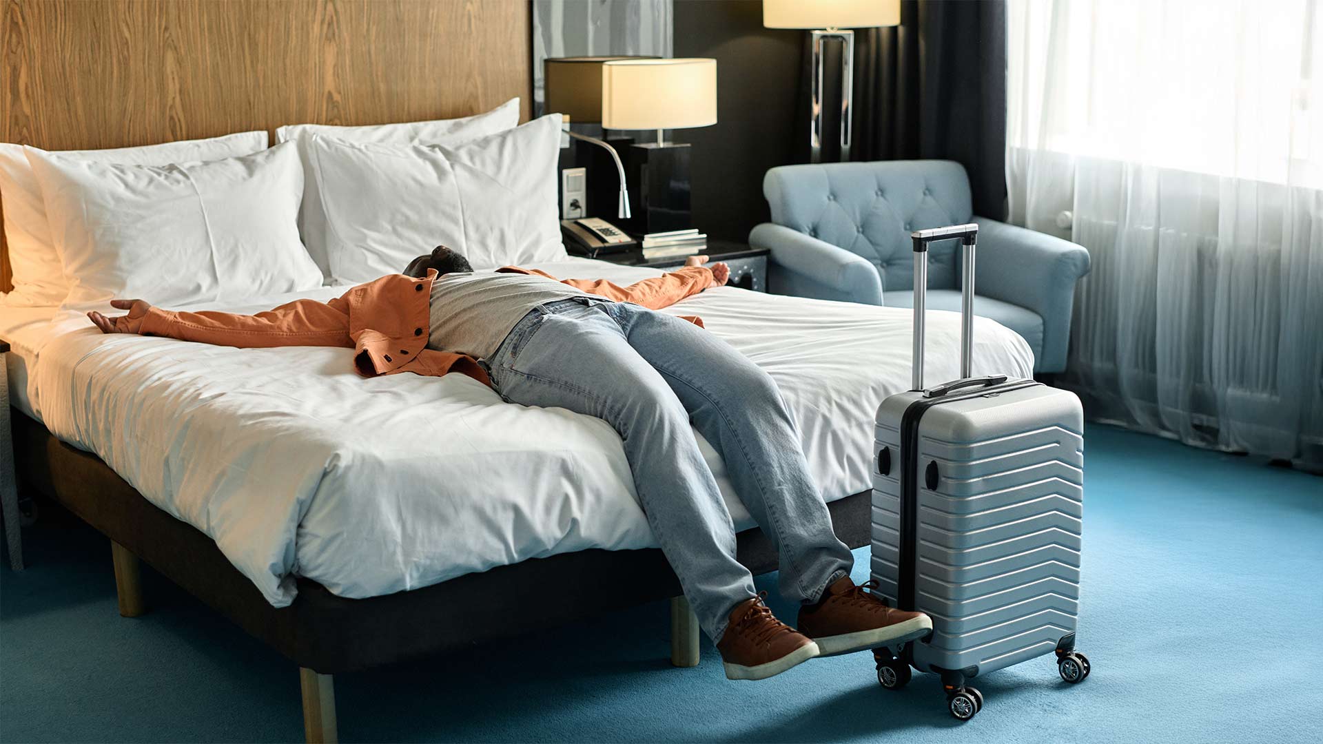 Exhausted traveler lying face-up on a hotel bed with suitcase nearby, in a cozy modern yet cheap hotel room.
